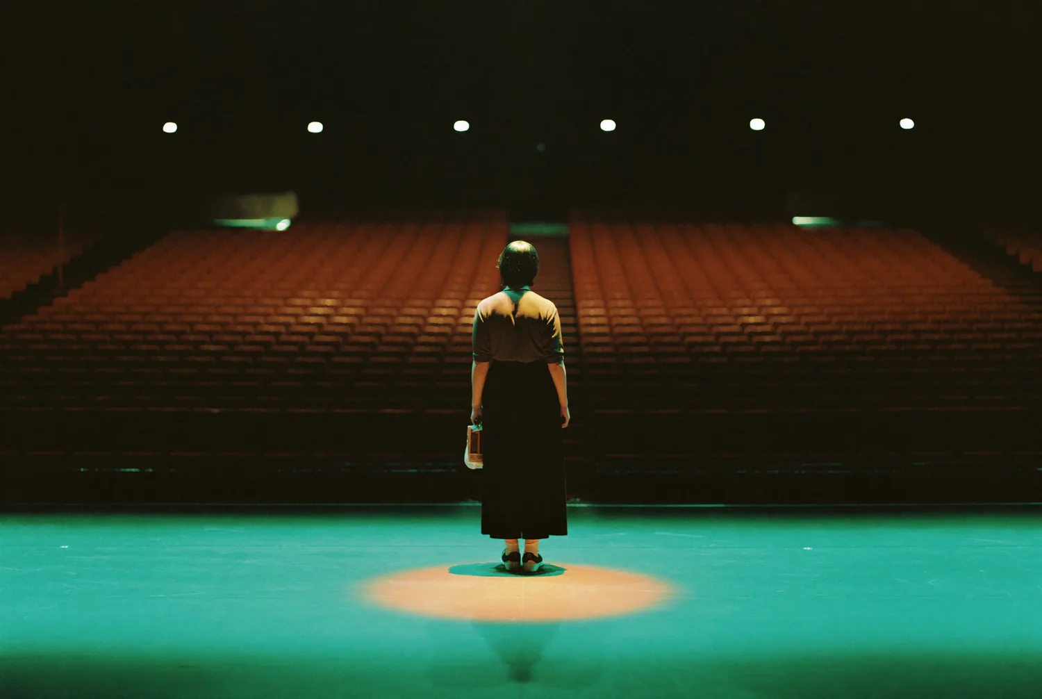 A lone figure under a single spotlight on a stage with empty seats beyond