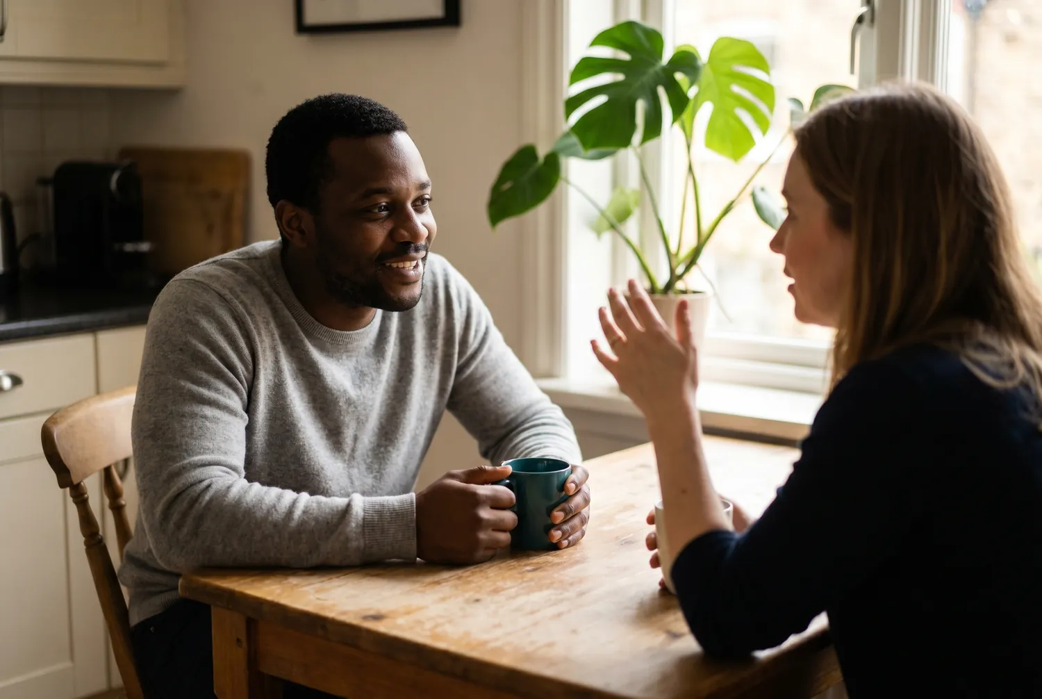 Two adults in genuine conversation at a sunlit kitchen table, one holding a deep teal mug